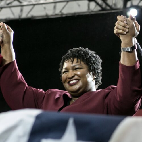 Stacey Abrams is smiling, standing behind a podium with flag draped over it. Her arms are in the air in a victory pose, holding hands with Barack Obama (not seen). She is wearing a burgundy colored suit, a watch, and a necklace.
