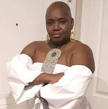 Cassandra Alexander-Green is wearing her bald, with gold hoop earrings, a large gold necklace, and a white off the shoulder long-sleeved top with her arms folded in front of her.