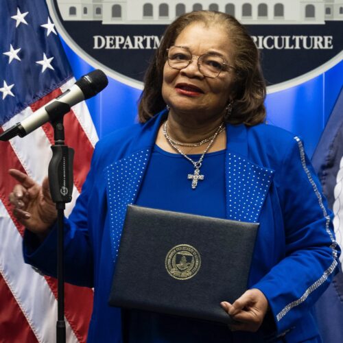 Alveda King, at her swearing in to office with the US Department of Agriculture. She is standing in front of a microphone, wearing glasses, a blue dress, and a cross. The US flag is on the left and the USDA seal is behind her.