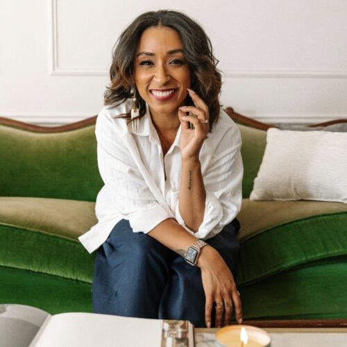 Nichelle Guidry smiling, wearing a white shirt, dark blue pants, a watch, and 2 rings. She is sitting on a green couch.