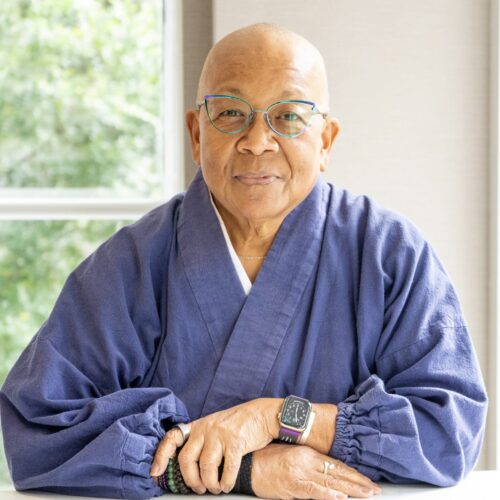 Myokei Caine-Barrett sitting, wearing glasses, a watch, and blue clothing, with a slight smile.