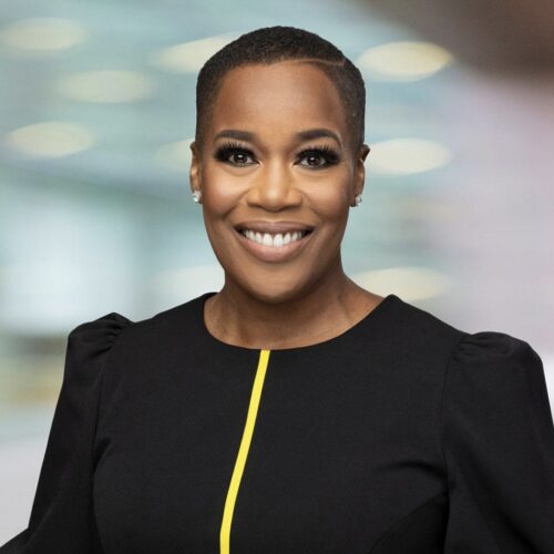 Saleemah Abdul-Ghafur smiling, wearing a black top with a yellow stripe down the center, and a soft pastel green background.