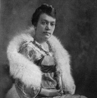 Coralie Franklin Cook sitting with her hands folded in her lap, wearing her hair short, with a dressy dress, a necklace, and a light-colored soft furry stole.