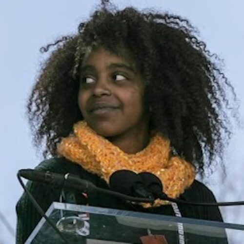 Naomi Wadler smiling, standing in front of a glass podium wearing a black sweater with an orange scarf and her hair in an Afro.