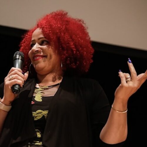Nikole Hannah-Jones smiling, holding a microphone, and the other hand in the air, wearing a black jacket with a black shirt and gold trim, red hair worn in an Afro.