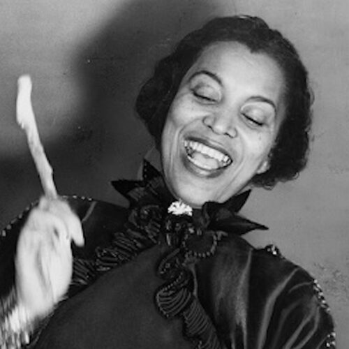 Zora Neale Hurston, smiling, holding a drumstick, beating a hountar, (mama drum), wearing short wavy hair, and a dress with a dark colored fancy high collared jacket and a brooch.