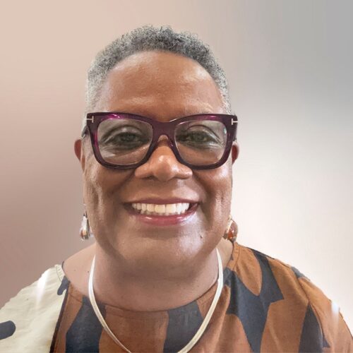 Renita Weems smiling, wearing short hair, glasses, earrings, a necklace, and a top with multiple shades of brown and cream colored design.