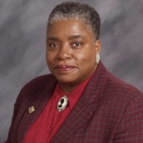 Cheryl Townsend Gilkes wearing short hair, earrings, burgundy jacket, red shirt, and brooch with a woman's silhouette.