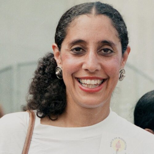 Carol Lani Guinier at the 30th Anniversary March on Washington. She is smiling, wearing her hair in a ponytail, with earrings, a white shirt with a 30th March sticker, and a purse over her shoulder.