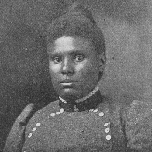 Emma B. Delaney wearing her hair up and a high-necked dress, puffed sleeves, a cross and a semi-circle of buttons. The photo is in black and white.
