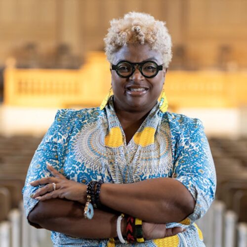 Melva Sampson, smiling and standing with her arms crossed, wearing black glasses, short blonde hair, long earrings, bracelets, a ring, and a blue patterned dress with yellow circles.