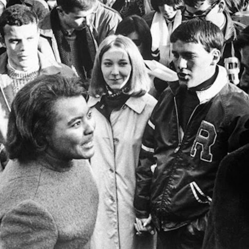 Prathia Hall speaking to someone with a crowd of young people in the background. She is wearing a sweater and her hair is shoulder length. The photo is in black and white.