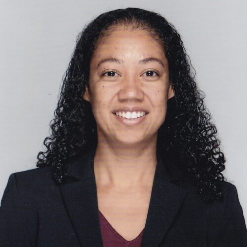 Rima Vesely-Flad smiling, wearing long curly hair, a black jacket, and a V-necked burgundy shirt.