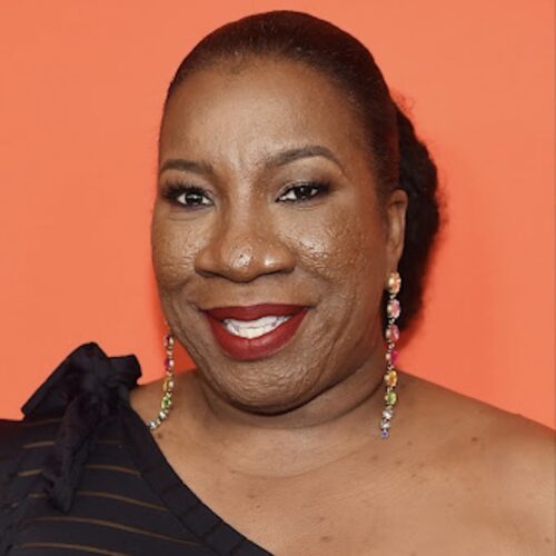 Tarana Burke smiling, wearing long earrings and a black fancy off-the-shoulder dress. The background behind her is a peachy orange color.