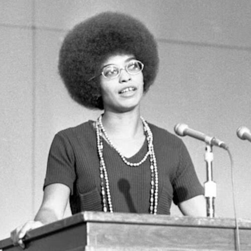 Angela Davis wearing a short-sleeved top with a long beaded necklace, her hair in an afro and glasses. She is standing in front of a podium with microphones. The photo is in black and white.