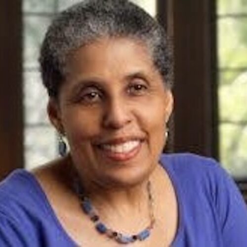 Barbara Smith smiling, wearing short black hair with gray highlights, earrings, necklace, and a bluish purple top.