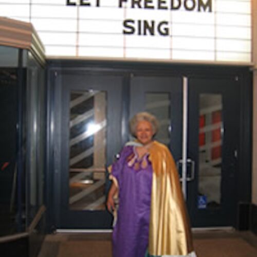 Carolivia Herron wearing short gray hear, with a purple gown and a gold shawl, standing in front of a theater marquee that says, “Let Freedom Ring.”