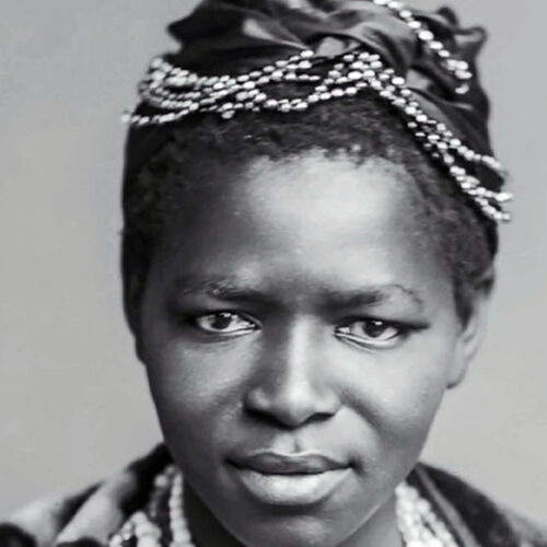Charlotte Maxeke close up, wearing her hair up in braids with decorative beads and a ribbon in her hair as well as a necklace. The photo is in black and white.