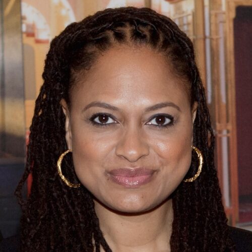 Ava DuVernay smiling, wearing long hair in locs, and gold hoop earrings.