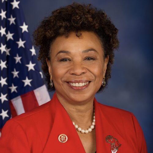 Barbara Lee smiling, wearing short curly hair, small hoop earrings, a pearl necklace, a red suit jacket with a gold Congressional pin and a gold pin in the outline of Africa. She is standing in front of the U.S. flag for her official Congressional photo with a royal blue background.