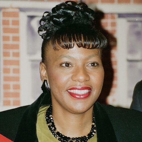 Dr. Bernice King smiling, wearing her hair in a curly updo with short curled bangs, earrings, a black and gold necklace, and a black jacket with a velvet collar over a lime green top. The background is a brick building.