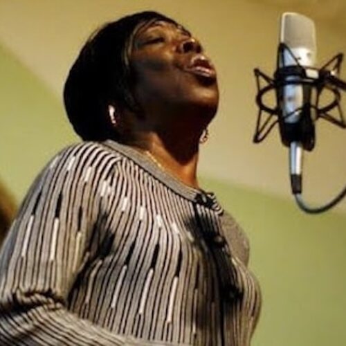 Betty Mae Fikes singing in front of a microphone, wearing short hair, earrings, and a top with thin vertical stripes in gray, black, and white.