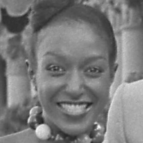Billie Blair smiling, with her hair in an updo and a large beaded choker necklace. The photo is in black and white.