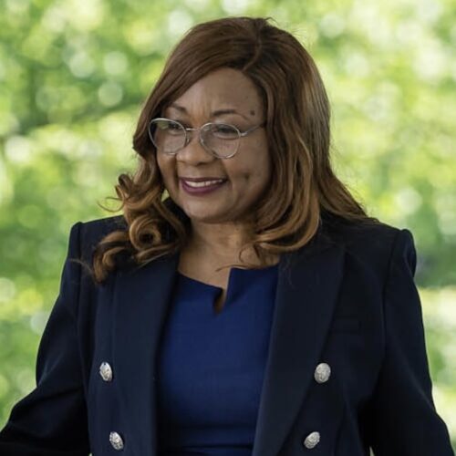 Catherine Coleman Flowers smiling, wearing shoulder-length light brown hair, glasses, and a royal blue jacket with silver buttons over a royal blue dress, with a blurred background of green leaves outside the White House.