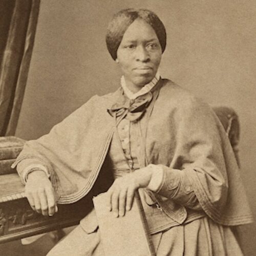 Amanda Berry Smith, seated, wearing her hair parted in the middle and pulled back, with a high-collared dress, a bow and lace at the collar, long-sleeved formal top with a cape, and a matching skirt. She is holding a book in her left hand and leaning on a desk with her right elbow. The photo is in sepia.