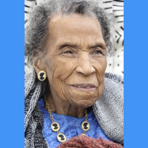 Amelia Boynton Robinson smiling, looking toward her left, wearing a blue top, a necklace and earrings with cameos of African women in black silhouettes, and a shawl.