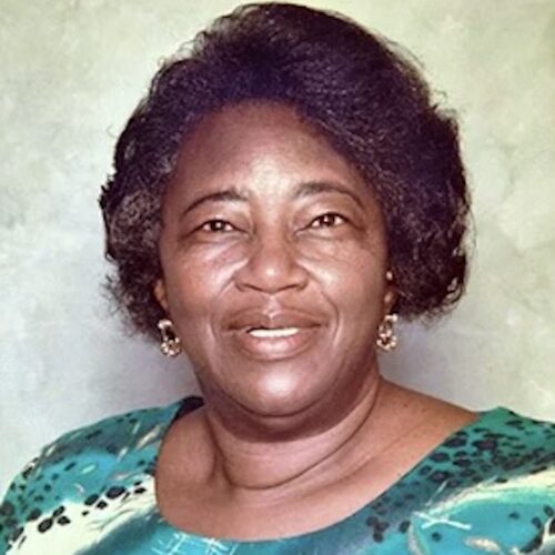 Catherine Stokes smiling, wearing short hair, gold earrings, and a green top with yellow and black patterned accents.