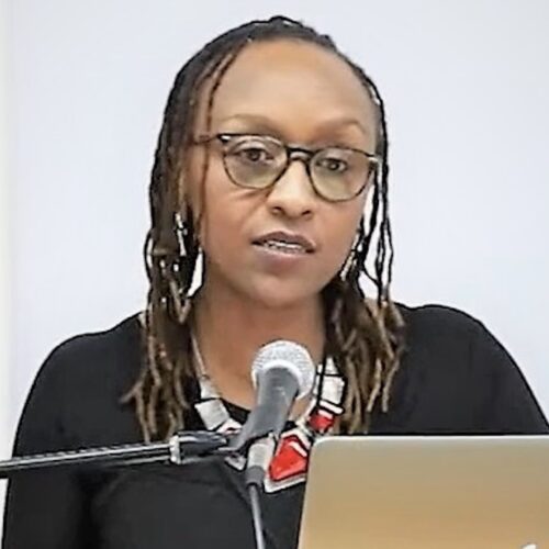 Elizabeth Mburu speaking in front of a microphone at a podium with her computer open. She is wearing her hair in long locs, glasses, a large white and red necklace, and a black top.