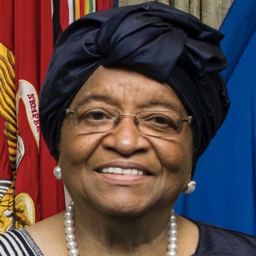 Liberian President Ellen Johnson Sirleaf, at an honor cordon hosted by Secretary of Defense Ash Carter at the Pentagon Feb. 25, 2015. She is smiling, wearing a dark blue head scarf and matching top, glasses, and pearl earrings and necklace, with flags behind her.