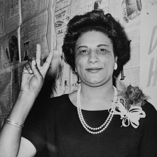 Constance Baker Motley with styled shoulder length hair, earrings, two strands of white bead or pearl necklaces, bracelets, and a corsage on her dark-colored top. Her right hand is raised in a peace sign. The photo is in black and white.