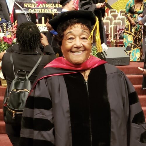 Alice Harris, smiling, receiving an Honorary Doctor of Humane Letters Degree in 2019. She is standing in a chapel, wearing a black academic robe with a red stole, a black cap with a gold tassel, and short curly hair.