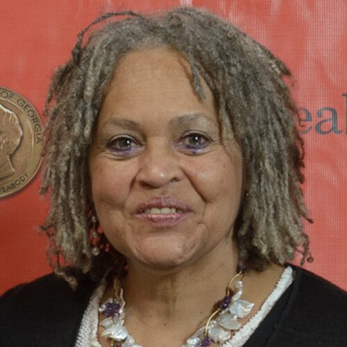 Charlene Hunter-Gault at the 73rd Peabody Awards, smiling, wearing shoulder-length locs, two shell and purple necklaces, and a black top with a white top underneath. The background is red.