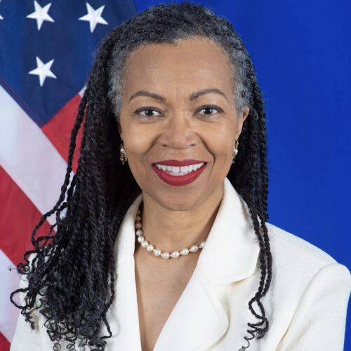 Gina Abercrombie-Winstanley, U.S. Ambassador, smiling, wearing long hair, red lipstick, pearl necklace, and a white suit jacket, in front of an American flag and royal blue background.