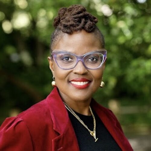 Angela Patton, smiling, wearing her hair up, with gold earrings and necklace, glasses, and a red suit with a black shirt. She is standing in front of trees.