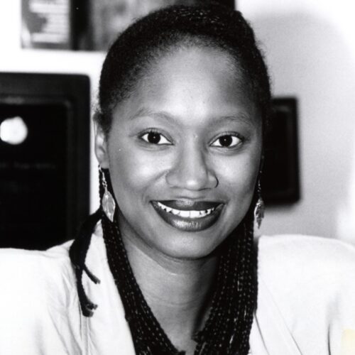 Elizabeth Brown-Guillory smiling, wearing her hair pulled back in long braids, as well as dangling leaf earrings, and a light-colored suit. The photo is in black and white.