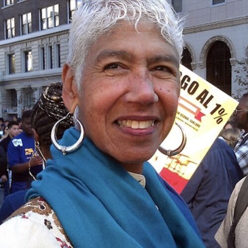 Ericka Huggins, smiling, wearing short gray hair, large hoop earrings, a white patterned top, and a blue scarf around her neck at the Occupy Oakland Protest on November 2, 2011.