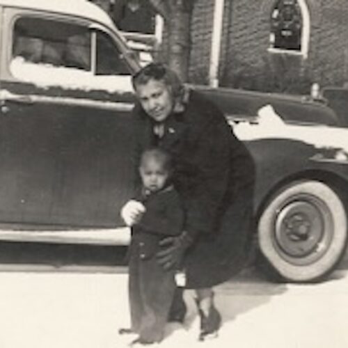 Eunice Jackson, standing in front of a church and a car in the snow, bending over and holding her very young son. Her hair is shoulder length, and she is wearing a dark top with a heavy overcoat, and snow boots. The photo is in sepia.