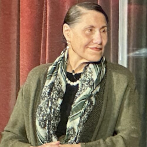 Evelyn Higginbotham, sitting on a panel, looking toward another speaker, with a slight smile, wearing her hair parted on her right side and pulled back, earrings, a white necklace, a microphone, and a black top under a dark green jacket with a patterned green scarf around her neck. Her hands are folded in front of her.