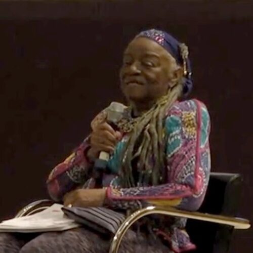 Faith Ringgold (born Faith Willi Jones) seated, holding a microphone, papers, and a purse. She is wearing her hair in long gray locs with a navy blue head scarf, and a patterned top, in pink, lavender, teal, and yellow.