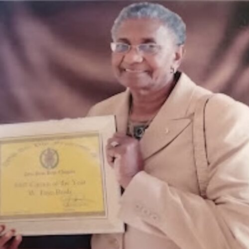 Faye Bush, smiling, sitting and holding her Citizen of the Year Award plaque. She is wearing short gray hair, glasses, gold earrings, a tan suit with a pin and a patterned tan and brown shirt underneath, and a gold ring and necklace.