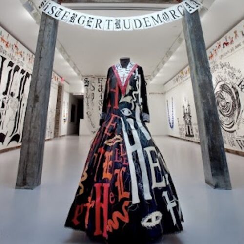 the installation of LESLEY DILL: HELL HELL HELL / HEAVEN HEAVEN HEAVEN : ENCOUNTERING SISTER GERTRUDE MORGAN & REVELATION, Arthur Roger Gallery in New Orleans, LA, 2010. It is a long dress with a V neck and cross. The left half of the dress vertically has Hell in red and orange letters. The right half of the dress says Heaven in white letters, with the dress in a black background. The letters Sister Gertrude Morgan are hung over the dress displayed in a grand hall with other artwork on the walls.