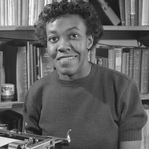 Gwendolyn Brooks in 1950, smiling, sitting in front of a typewriter, wearing short curly hair, sweater, skirt, bracelet, and wedding rings.