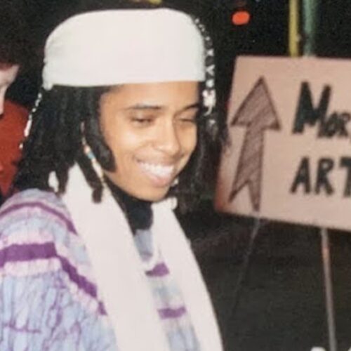 Gwynelle Dismukes smiling, wearing a white head scarf, shoulder-length locs, a white scarf around her neck, and a tye-died light blue, purple, and white top with slender purple veins of color. In the background is a sign with an arrow pointing to More Art.