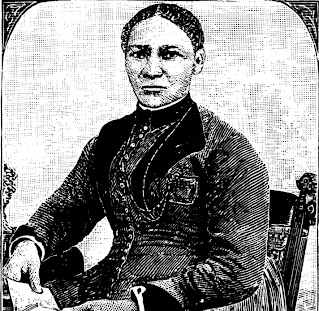 Harriet Baker, wearing her hair parted and pulled back, and a dark colored long sleeved dress with a high collar and buttons down the front. She is seated holding an open book with both hands. The photo is in black and white.